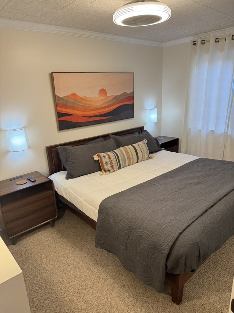 Master suite with a king bed, sunset artwork above the headboard, matching bedside lamps and sheer white curtains