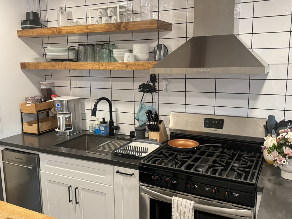Cottage kitchen with gas range, stainless hood, black countertops and open wood shelving stocked with dishes