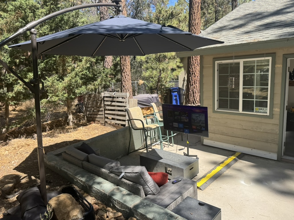 Back patio with a large sectional sofa, gas fire table, cantilever umbrella and an outdoor television among the pines