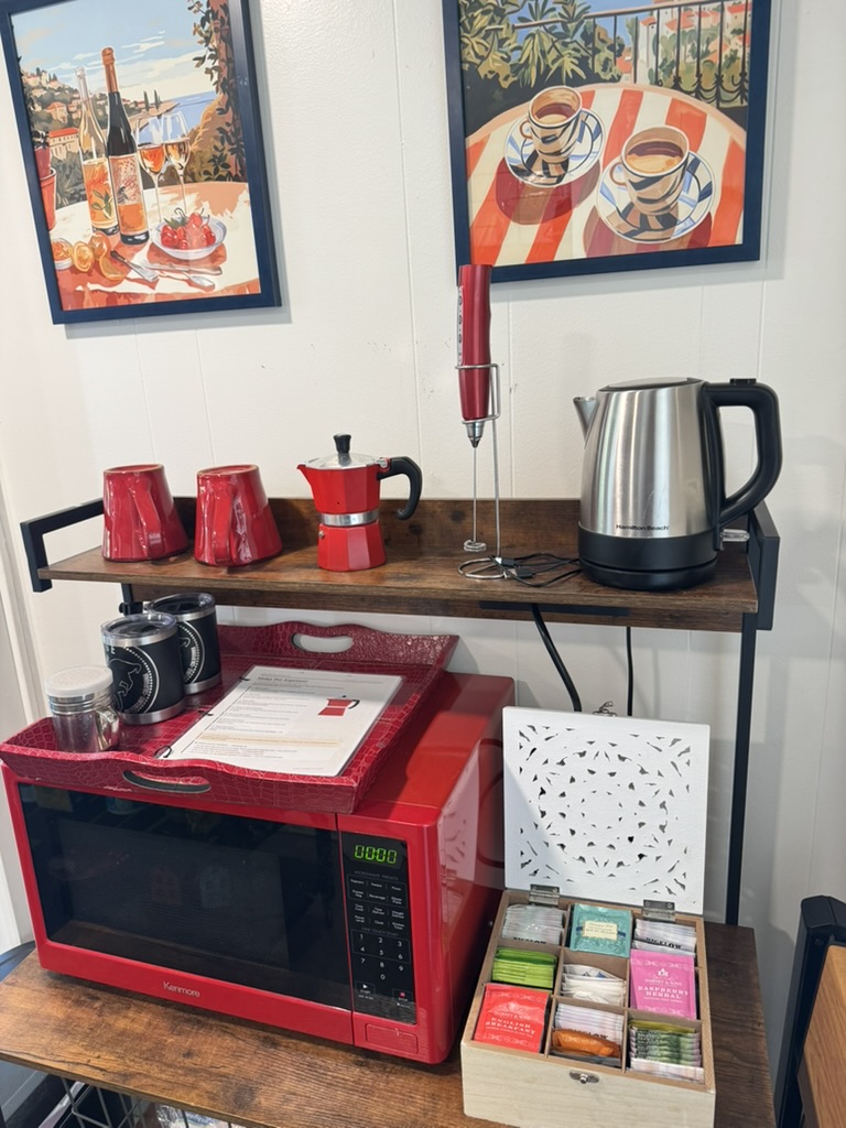 Coffee and tea station with a moka pot, red mugs, electric kettle, milk frother, red microwave and a boxed tea assortment